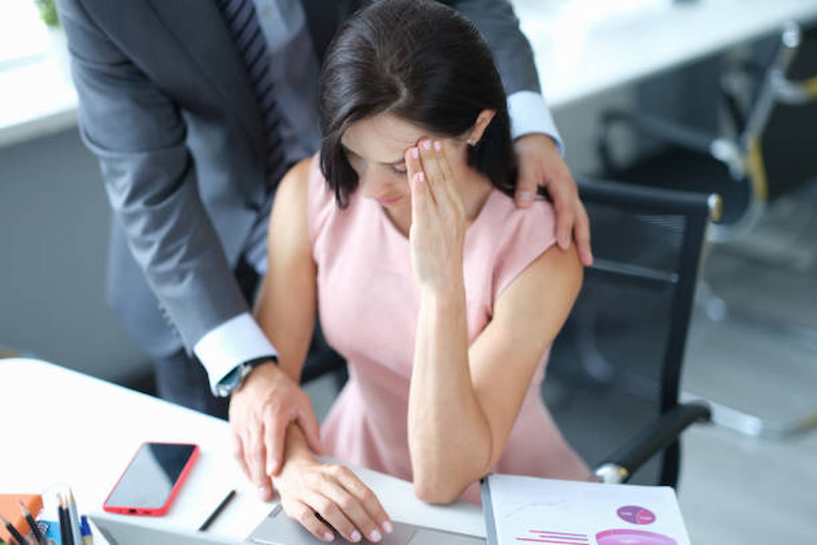 A woman sits at a desk, looking stressed, while a man's hands rest on her shoulder and arm. A phone and documents scatter the surface. Perhaps it's time to consult an employment lawyer in Los Angeles for guidance and support. Employee Rights>Sexual Harassment