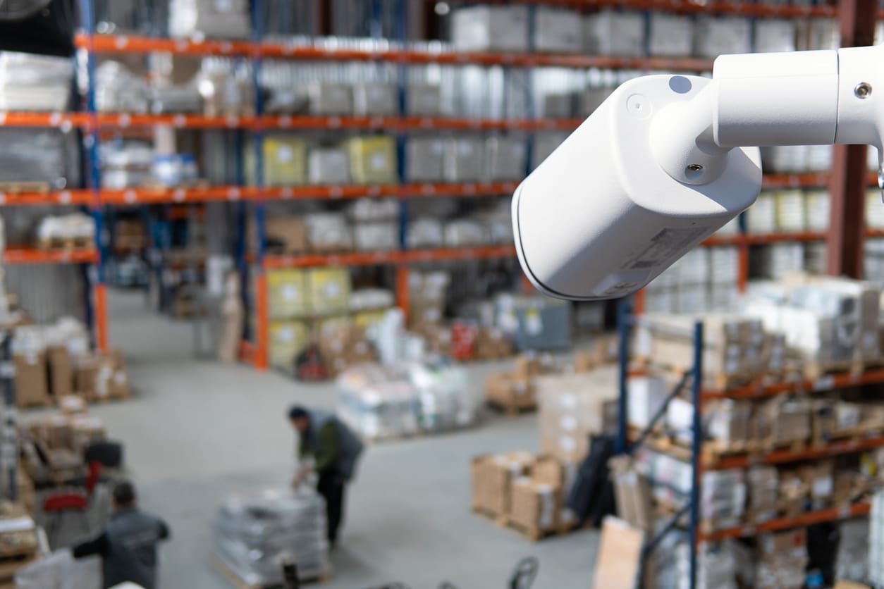A security camera overlooks a bustling warehouse with shelves full of boxes, capturing workers diligently handling tasks. In the backdrop, an employment lawyer from Los Angeles could be advocating for fair workplace practices amidst this hive of activity. Employee Rights