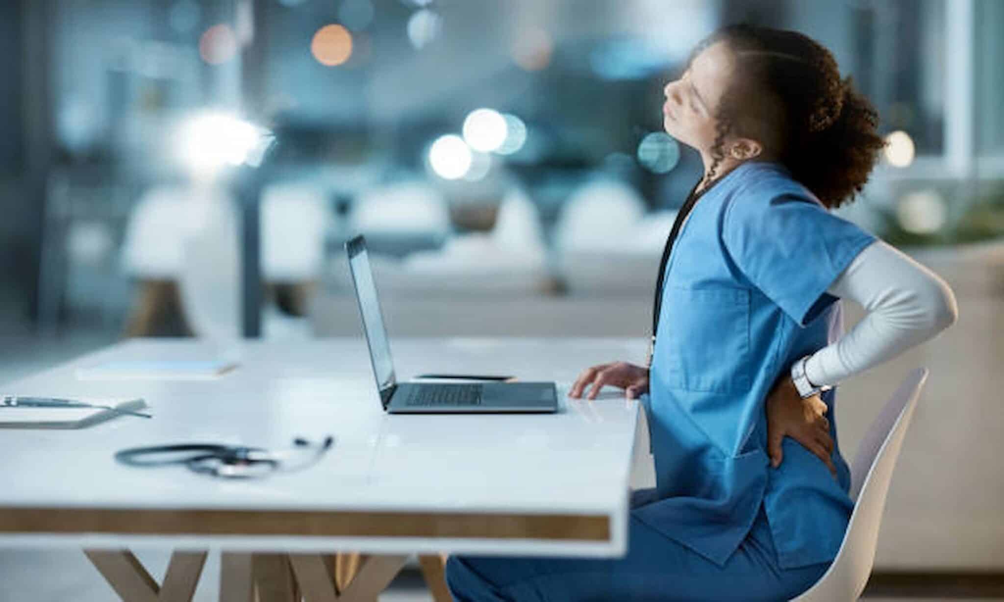 A person in blue scrubs sits at a desk with a laptop, stretching and holding their lower back, suggesting discomfort. Nearby is a brochure for an employment lawyer in Los Angeles, hinting at the stress and complexity of workplace issues that may require legal counsel. Employee Rights