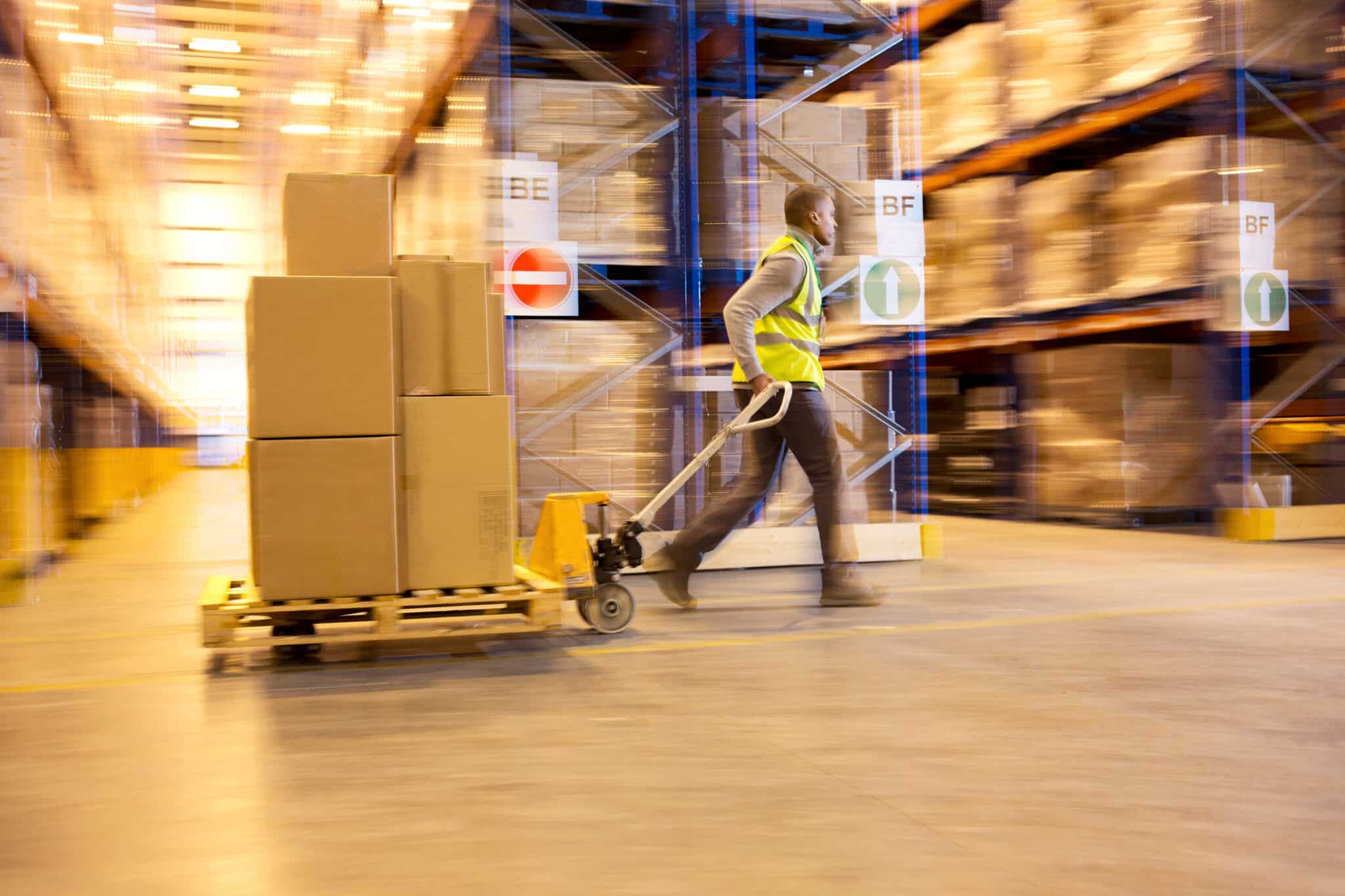 A warehouse worker in a high-visibility vest pulls a pallet jack with stacked boxes through an aisle of shelves filled with packages, ensuring safe operations for all employees, aligning with the guidelines you might expect from a diligent Los Angeles employment attorney. Employee Rights
