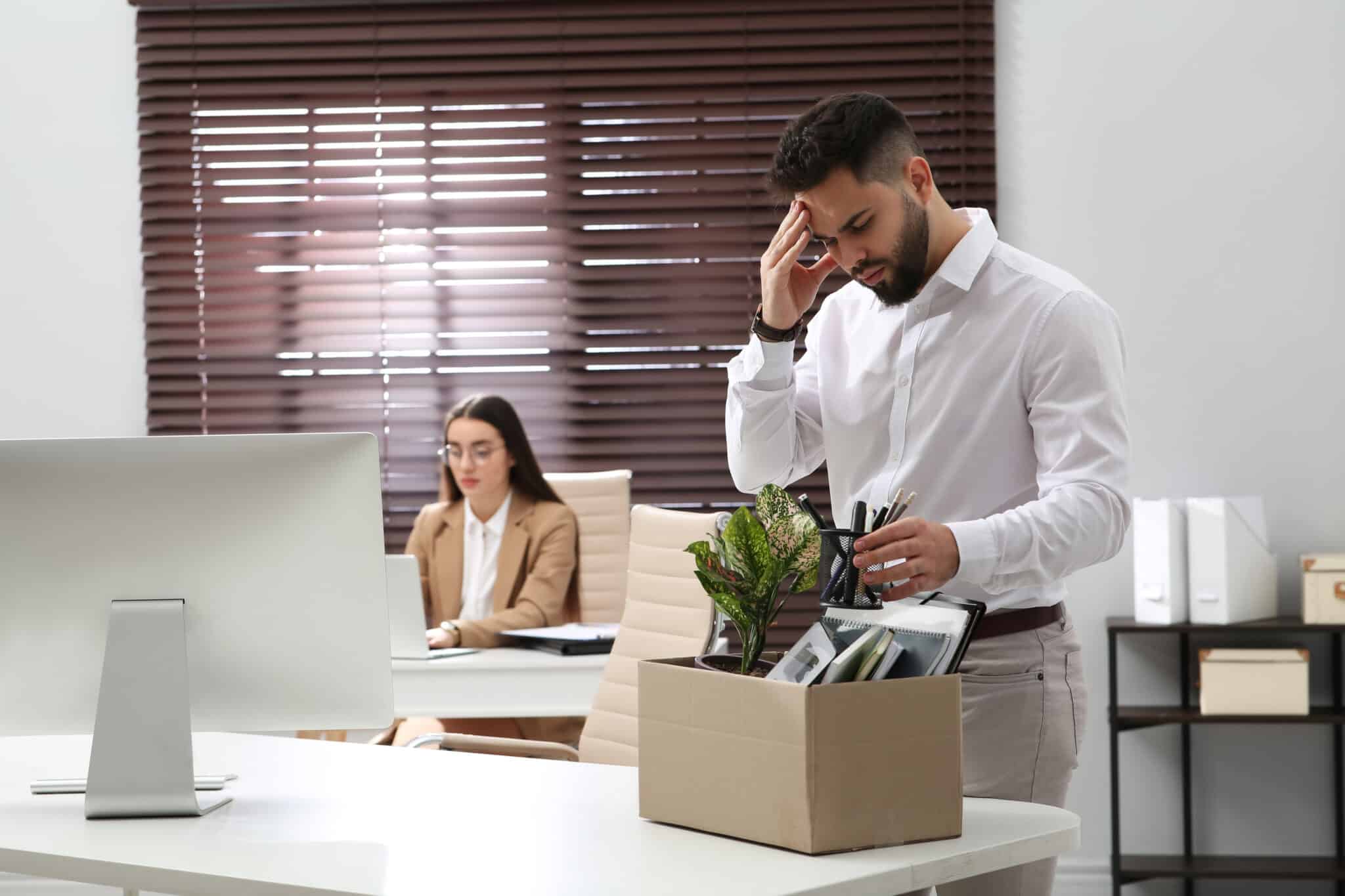 A man stands in an office with a box of personal belongings, looking concerned. In the background, a woman works diligently at her desk. If facing workplace challenges, consulting an employment lawyer in Los Angeles might offer clarity and protection in uncertain times. Employee Rights>Wrongful Termination
