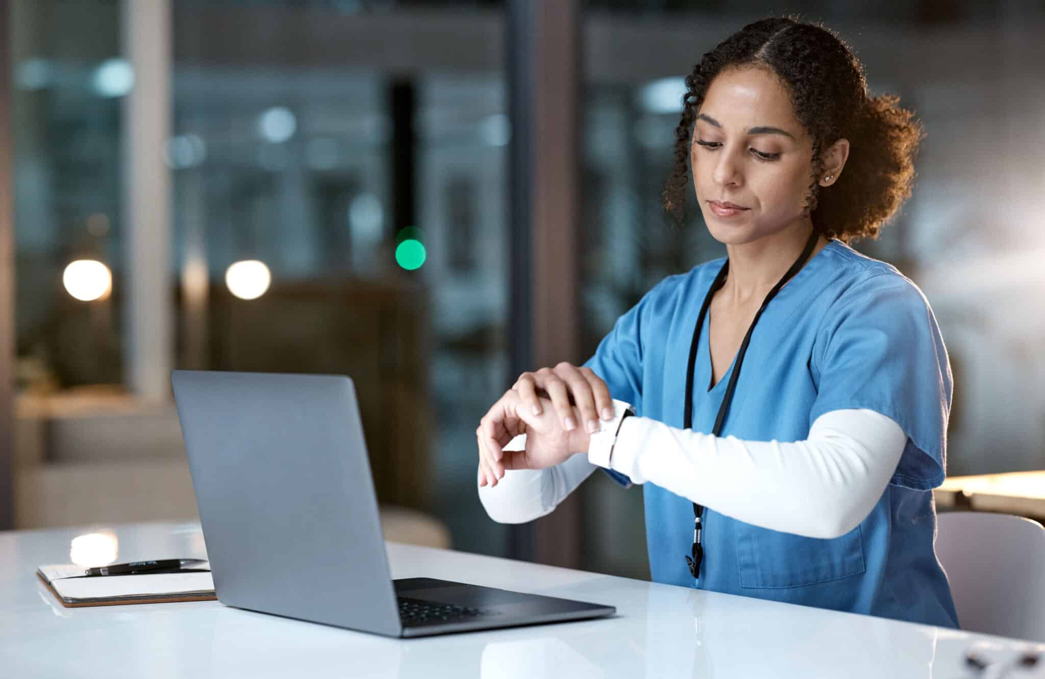 A woman in medical scrubs sits at a laptop, checking her smartwatch with a clipboard and pen nearby in the dimly lit room. Despite her medical career, she's contemplating consultations with an employment lawyer in Los Angeles to explore her rights within the healthcare industry. Employment Law