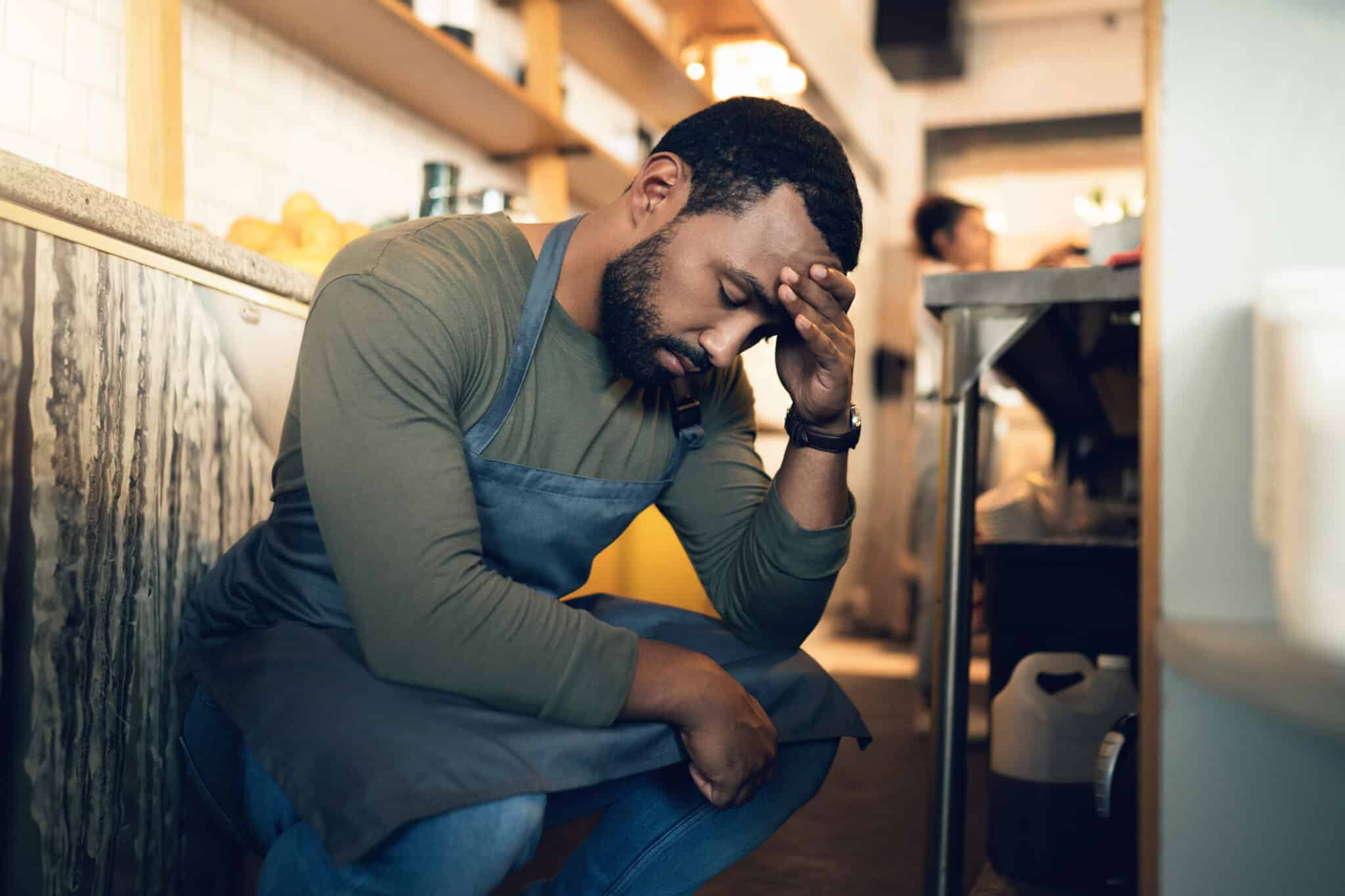 Man in an apron crouching in a kitchen, resting his head on his hand, appears thoughtful or stressed, perhaps contemplating calling an employment lawyer in Los Angeles for advice. Employment Law