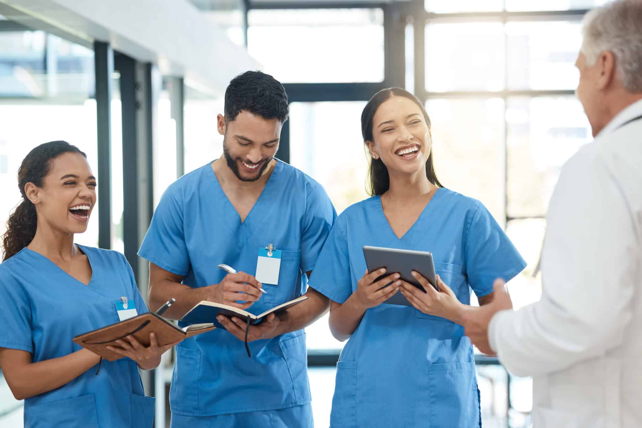 Four healthcare professionals in blue scrubs are smiling and talking in a hallway, resembling the collaborative spirit of an employment lawyer in Los Angeles, while holding notebooks and tablets. Employee Rights>Wage and Hour