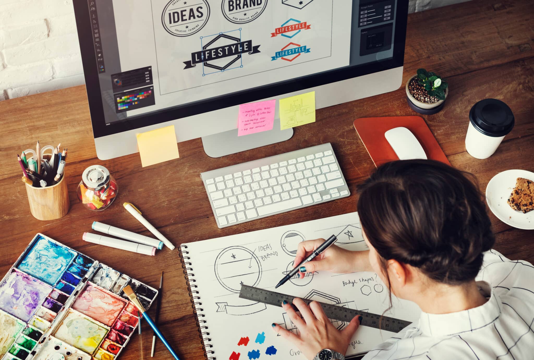 A person is sketching design ideas with markers and a ruler at a desk, which also has a computer displaying branding concepts, colorful markers, and a cup. Nearby, notes from a Los Angeles employment attorney are tucked under the keyboard, inspiring workplace policy designs. Employment Law