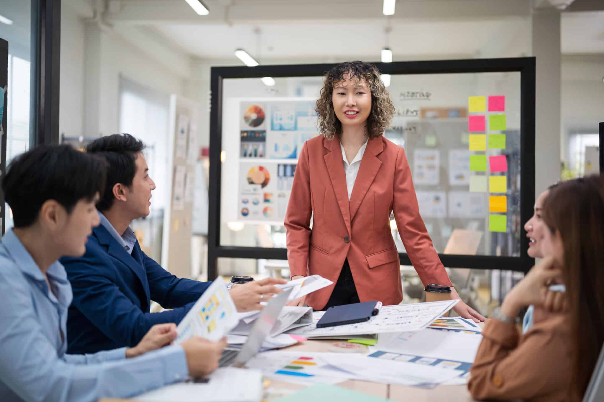A woman in a business suit speaks at a conference table with four colleagues, papers and charts spread across the table in a brightly lit office. An experienced Los Angeles employment attorney, she confidently addresses the team on effective workplace strategies. Employee Rights