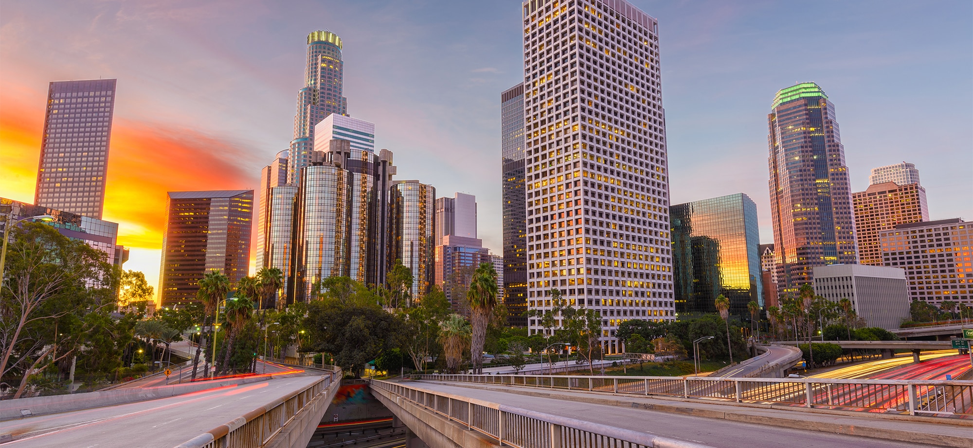 A city skyline at sunset with towering skyscrapers and a multi-lane road in the foreground, surrounded by trees, embodies the dynamic pace of Los Angeles. Just like an employment lawyer in Los Angeles, it stands as a symbol of relentless ambition and steadfast support amidst the bustling landscape.